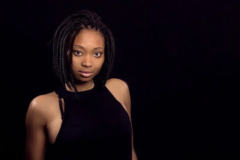 A woman with long braids in a black halter top, lit against a black ground