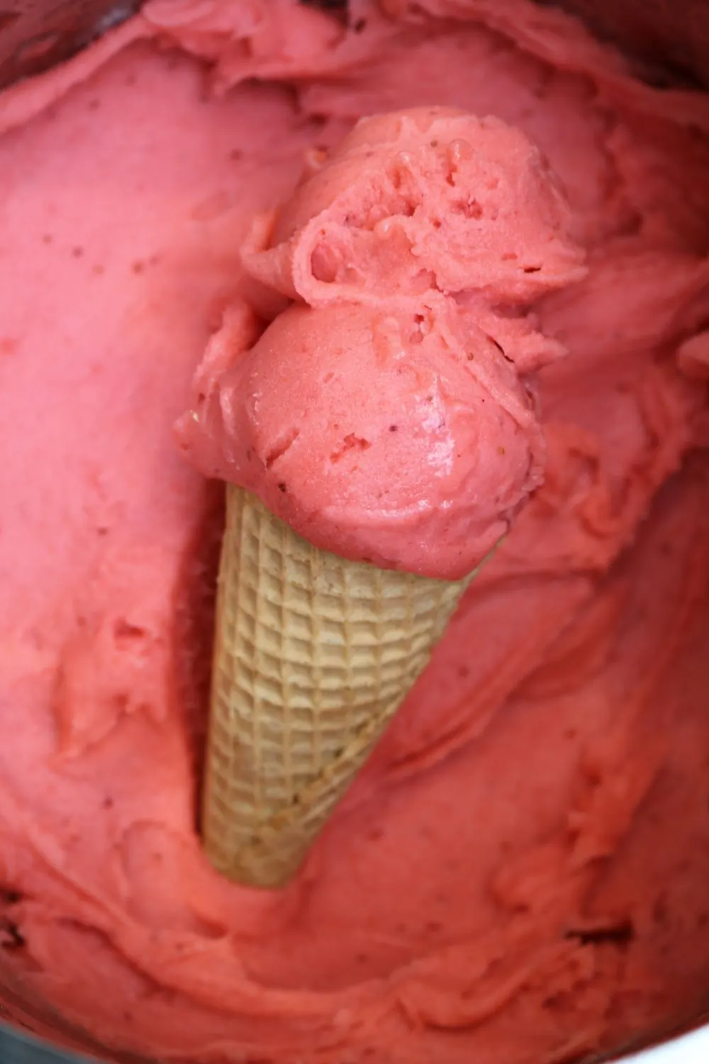 A pink strawberry scoop in a waffle cone held up against a bright sky
