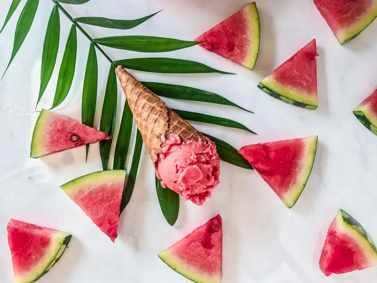 A pink watermelon sorbet in a waffle cone laid on white marble between palm fronds and cut watermelon wedges