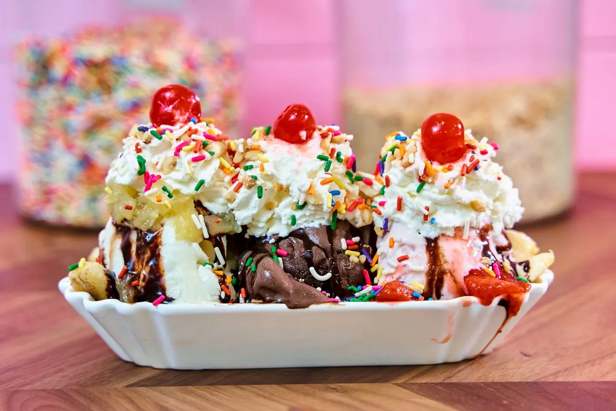 A banana split in a long white dish under whipped cream, rainbow sprinkles and three glacé cherries, jars of toppings behind it