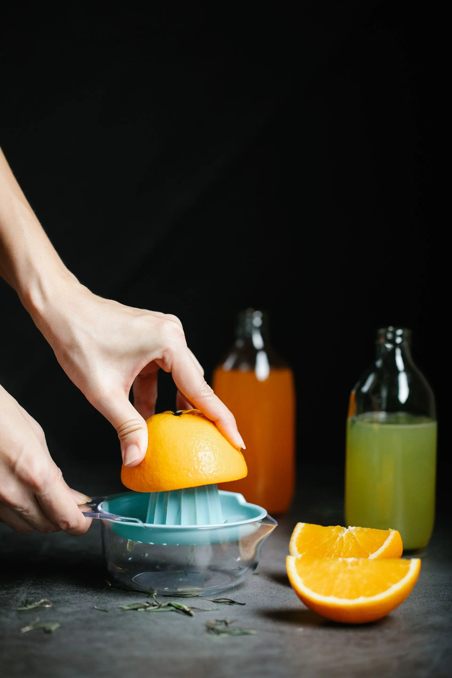 Hands pressing a halved orange over a juicer, filled bottles waiting on the dark bench behind