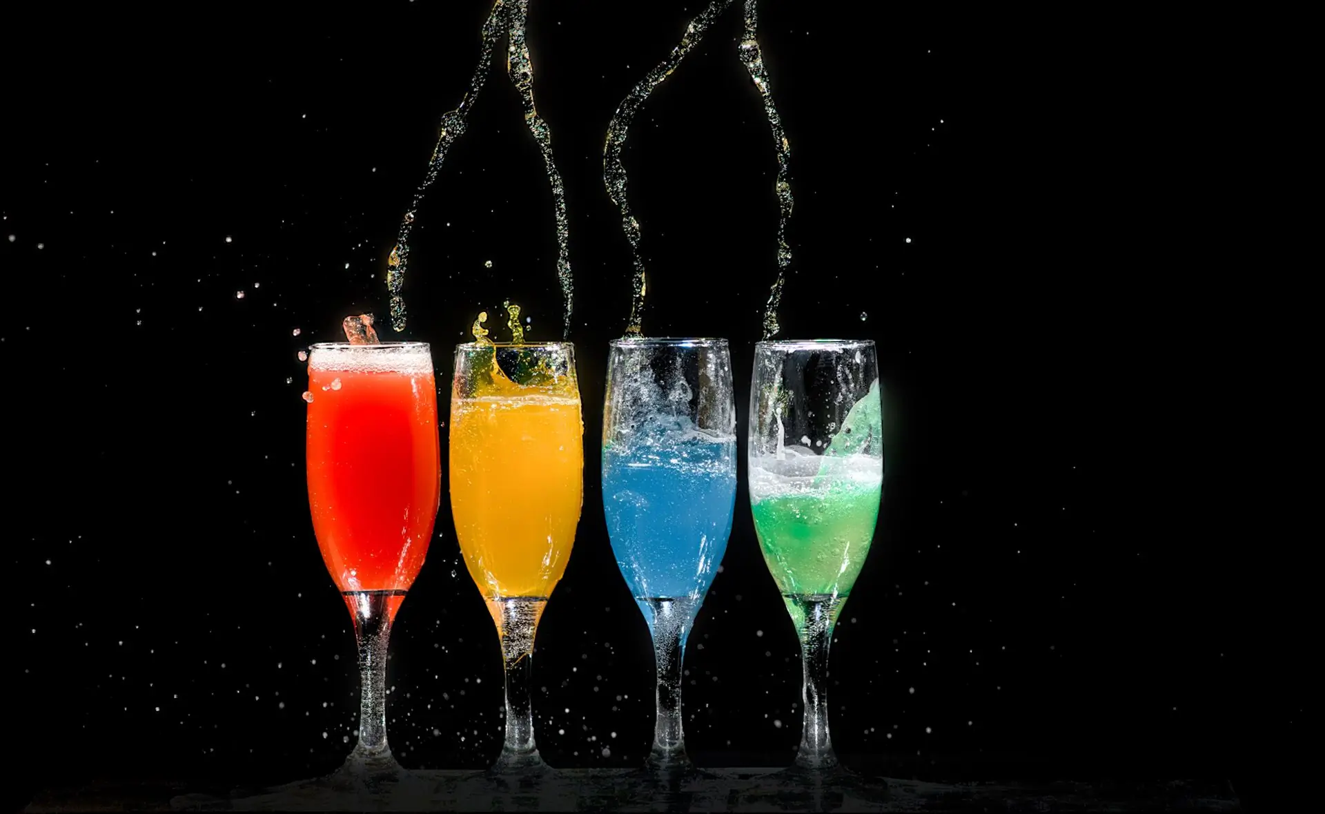 Four tall glasses of red, orange, blue and green fruit drinks with pour streams frozen mid air on a black ground