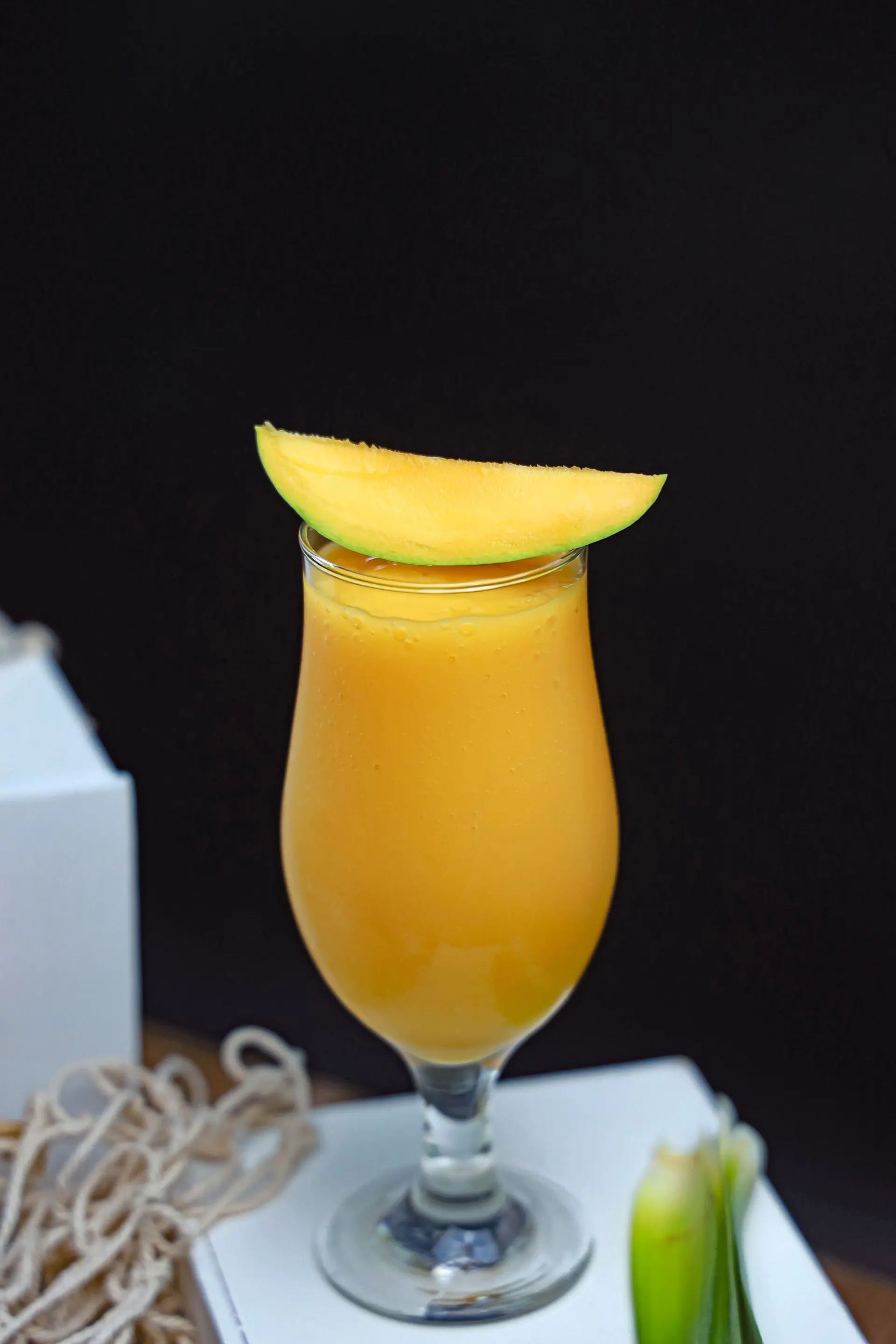 A tall glass of mango juice with fresh mango halves beside it on a warm amber ground