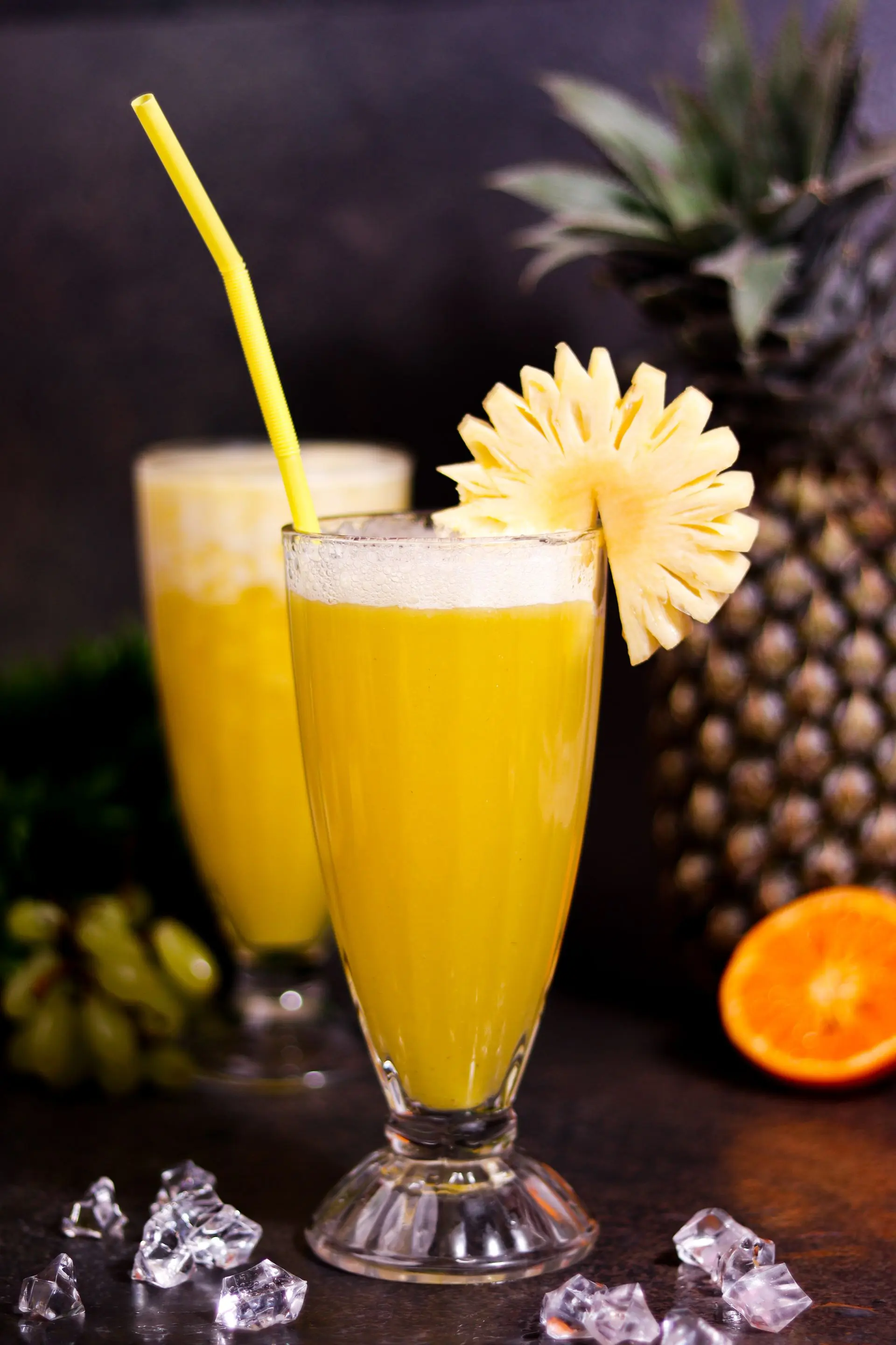 A glass of pineapple drink with a wedge of pineapple on the rim against a yellow ground