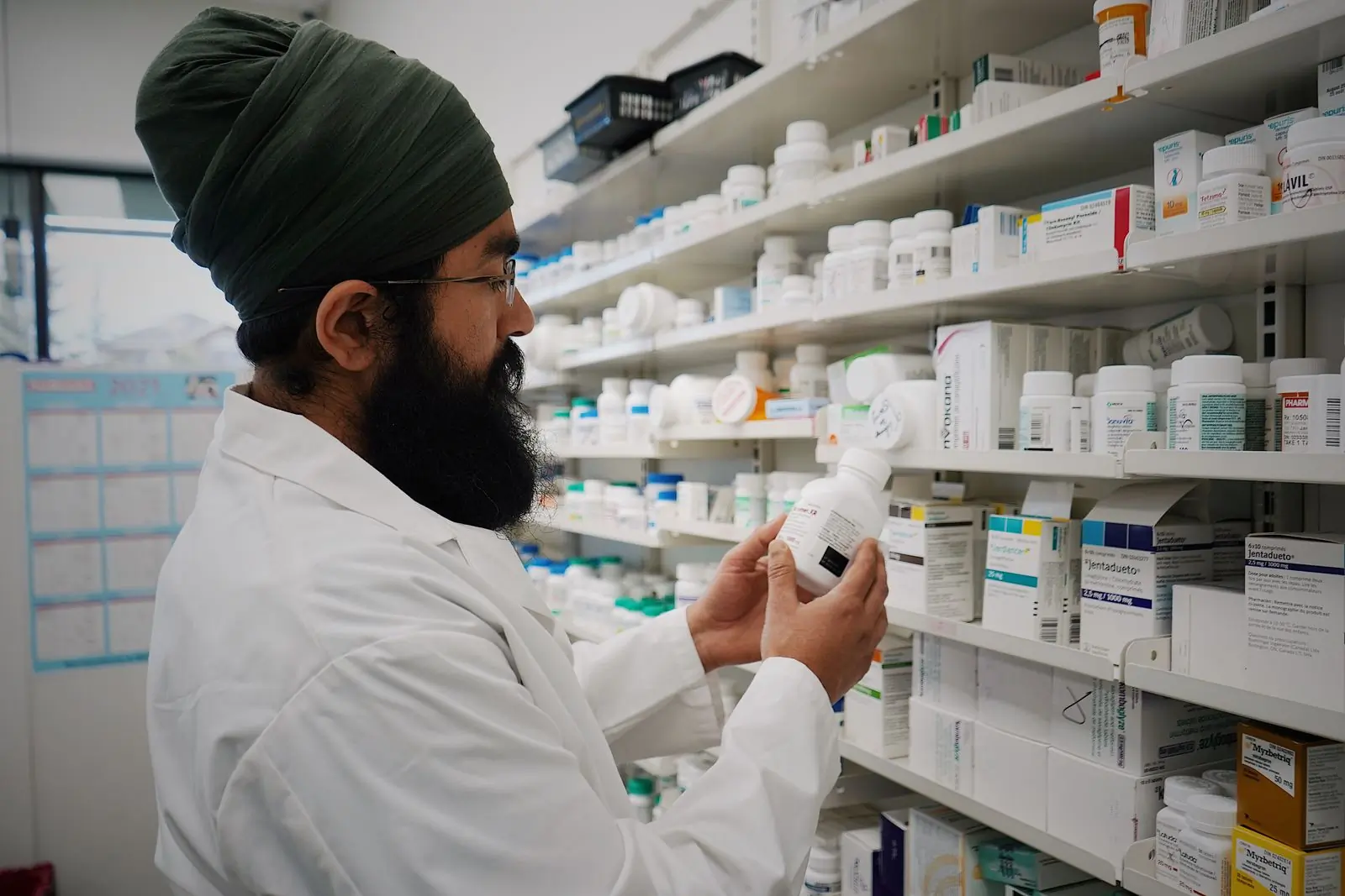 A pharmacist in a white coat and a patterned turban holding a supplement bottle up to read its label in front of the dispensary shelves