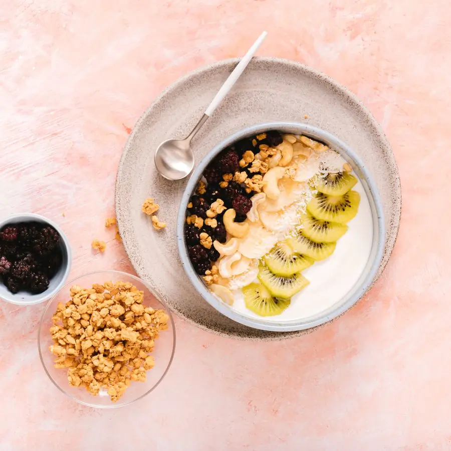 A bowl of yoghurt topped with sliced kiwi, cashews, blackberries and granola, seen from above