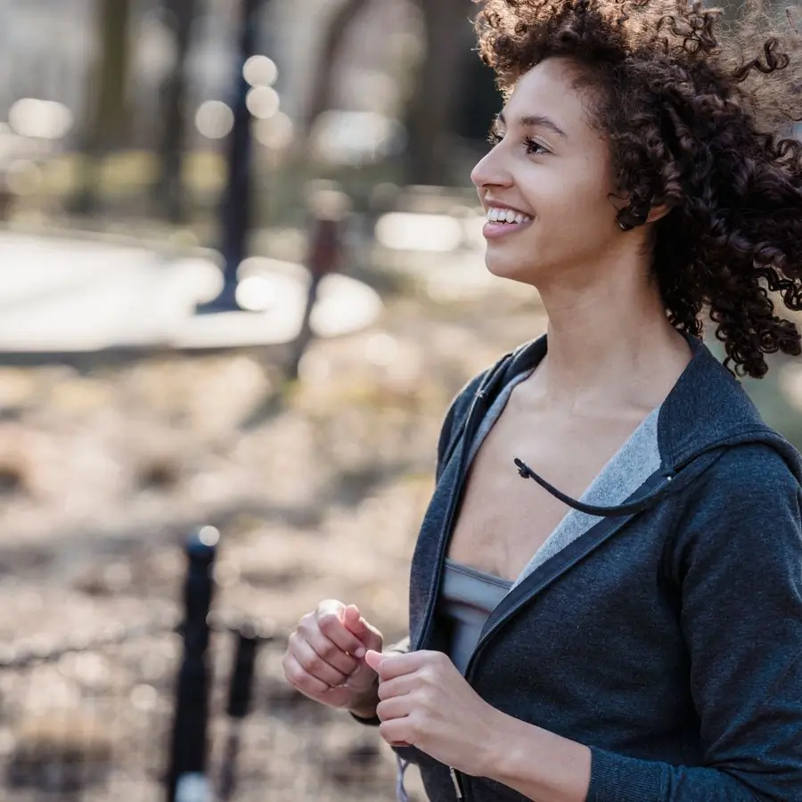 A smiling woman with curly hair running through a sunlit park in a grey zip-up jacket