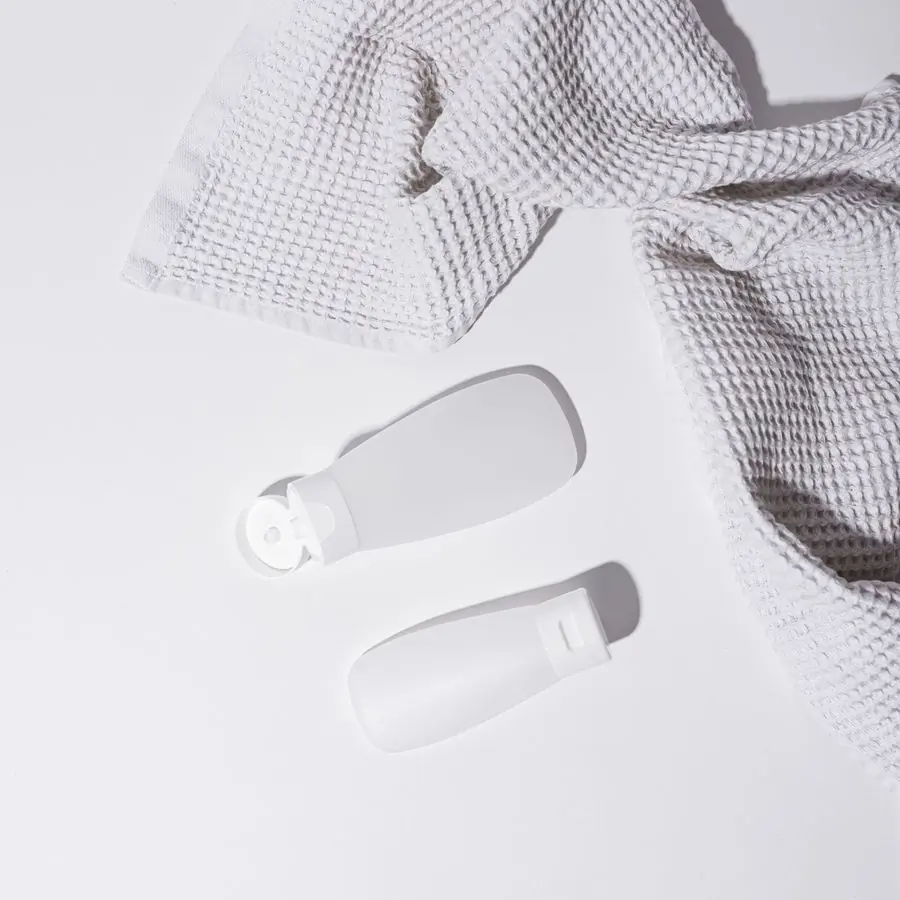 Two plain white cosmetic bottles on a white surface beside a crumpled waffle-weave towel