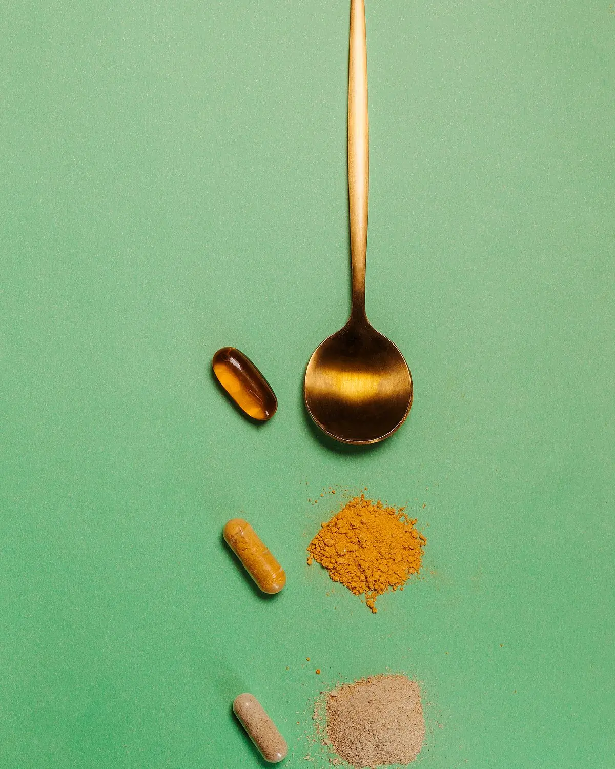 A golden spoon on a green surface above small piles of turmeric and beige capsule powder