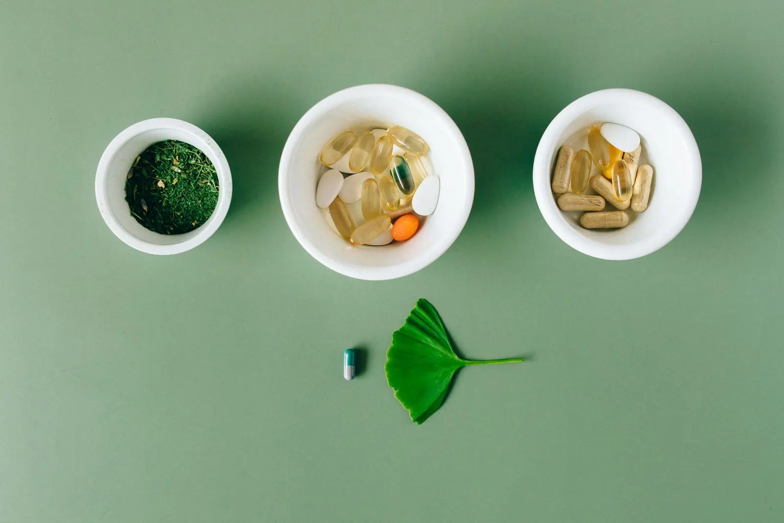 Three white bowls of capsules, tablets and green powder on a soft sage ground with a ginkgo leaf