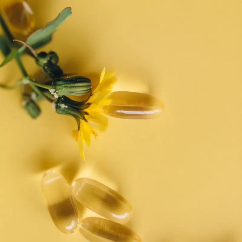 Golden softgel capsules scattered on a yellow surface beside a small dandelion flower