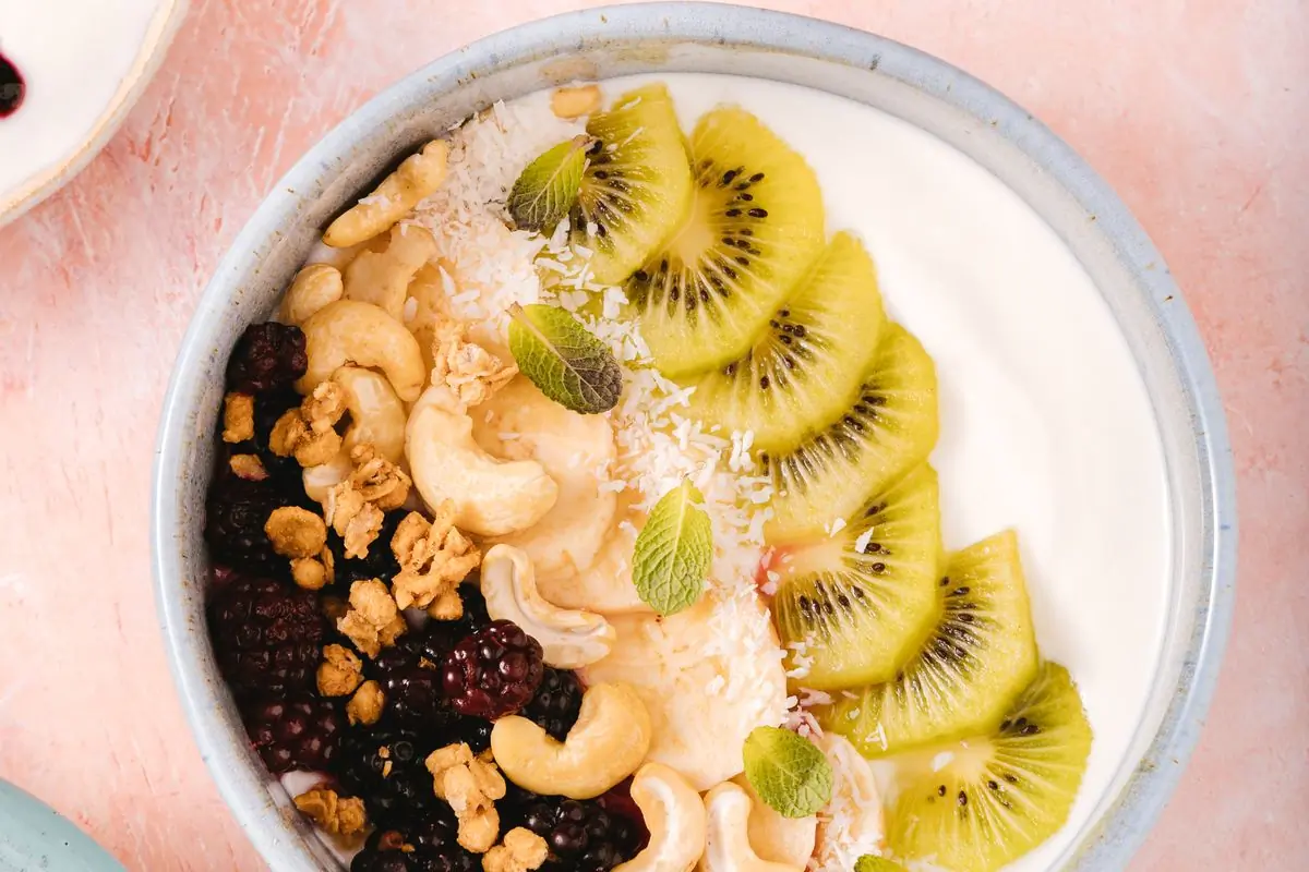 A yoghurt bowl topped with kiwi, blackberries, cashews and granola on a pale surface