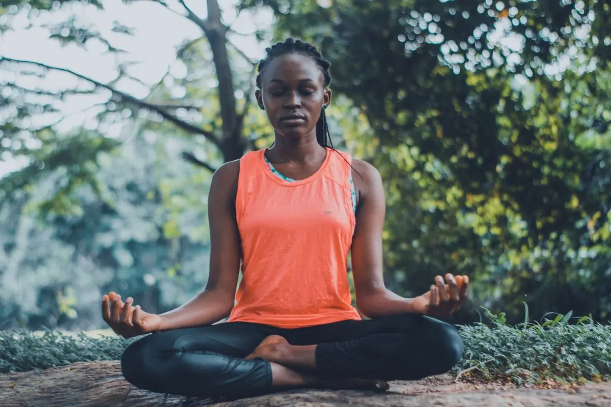 A woman sitting cross-legged in a meditation pose on a rock surrounded by green foliage