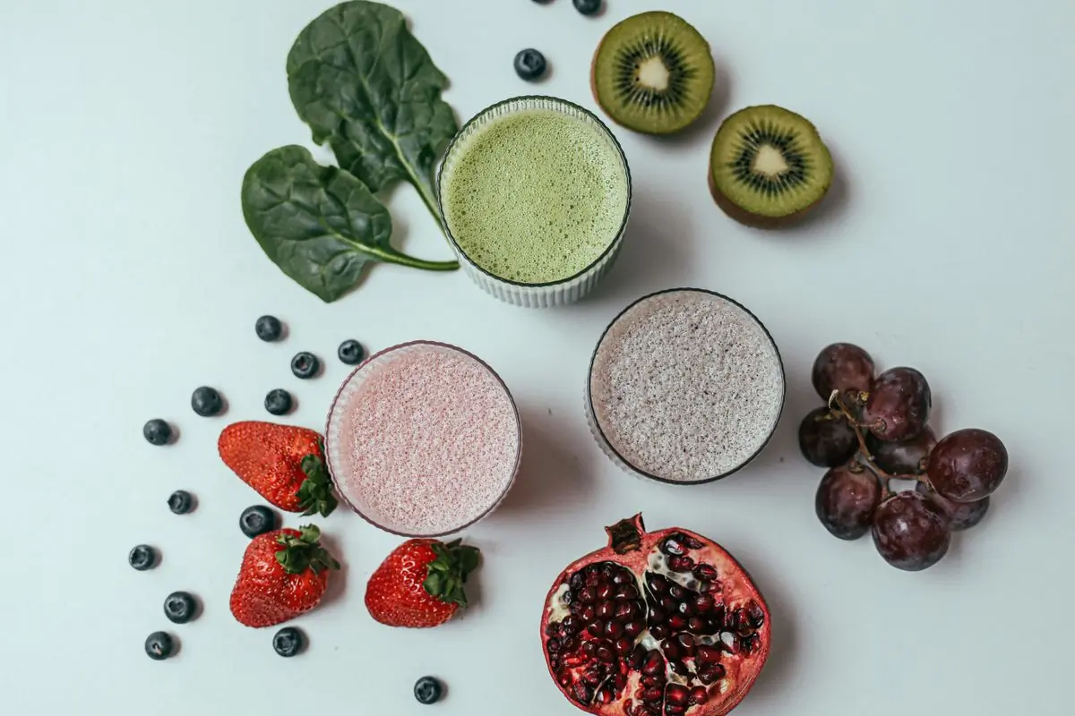 Three smoothies on a white surface surrounded by spinach, kiwi, berries, grapes and pomegranate