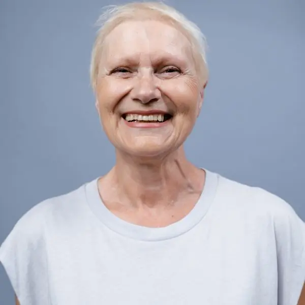 An older woman in a white t-shirt laughing warmly against a soft blue-grey backdrop