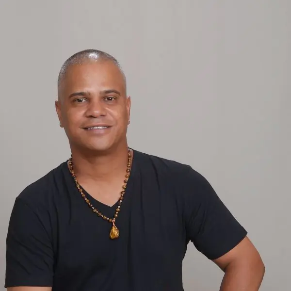 A smiling man in a black v-neck and beaded necklace against a plain grey background
