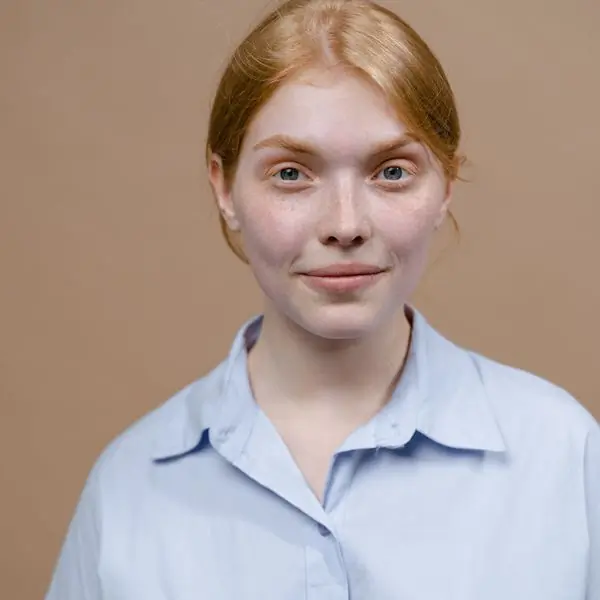 A young woman with red hair in a pale blue shirt, photographed against a warm beige backdrop