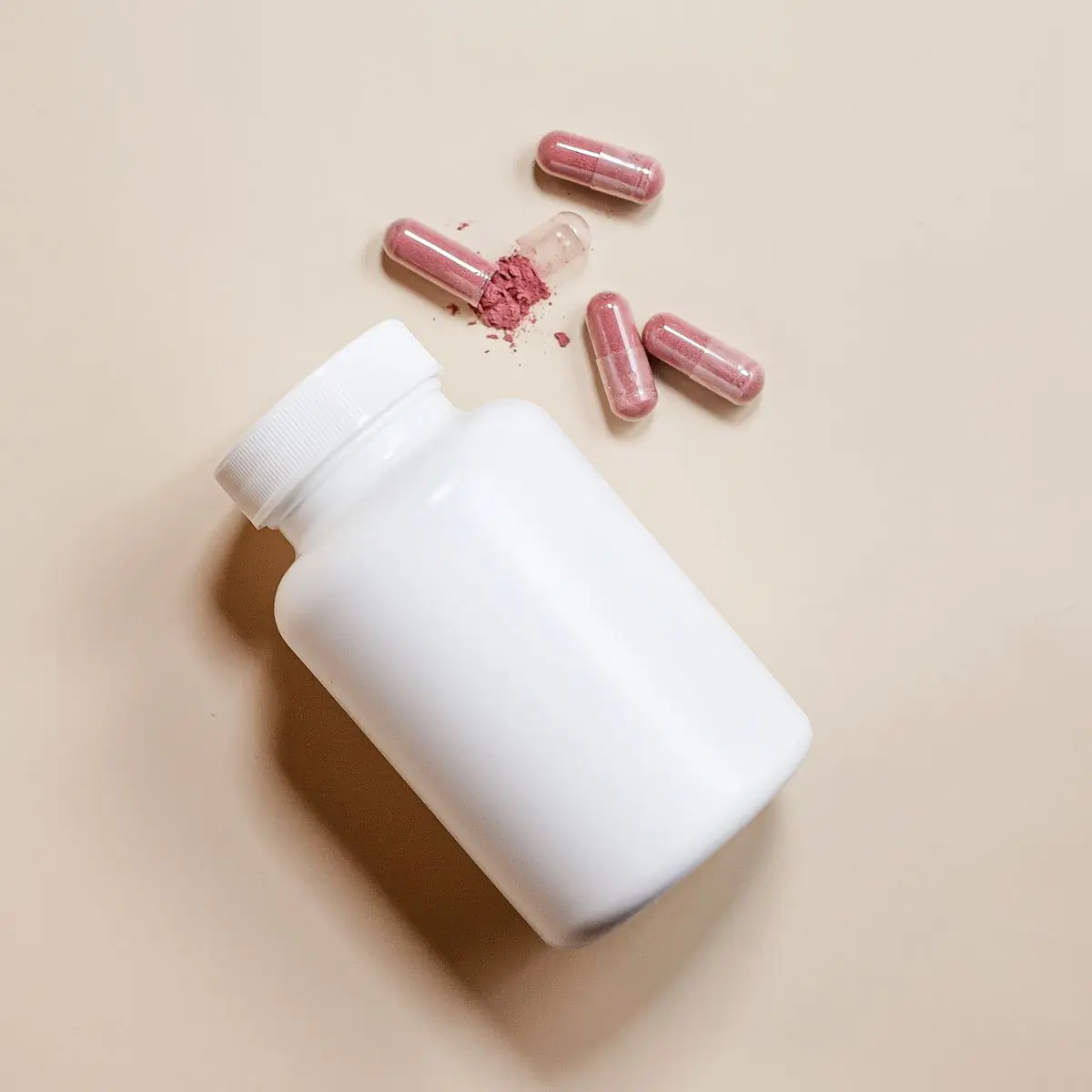 A white supplement bottle seen from above on beige, with capsules and a dusting of powder above it