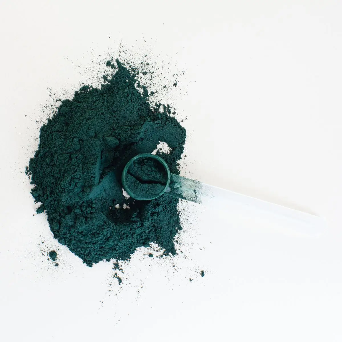 A mound of deep green spirulina powder on white with a clear measuring scoop resting in it