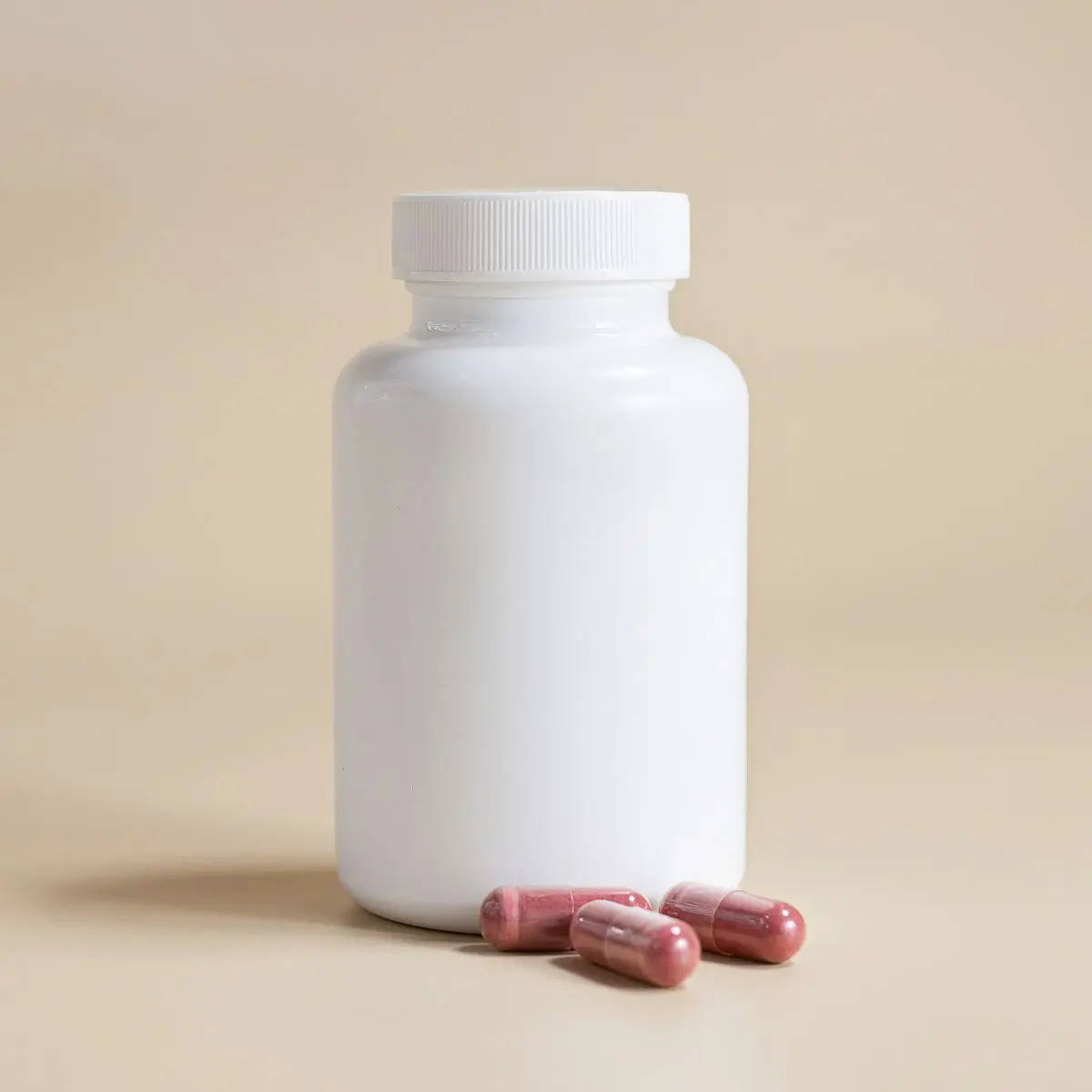 A white supplement bottle standing on a beige ground with three capsules resting at its base