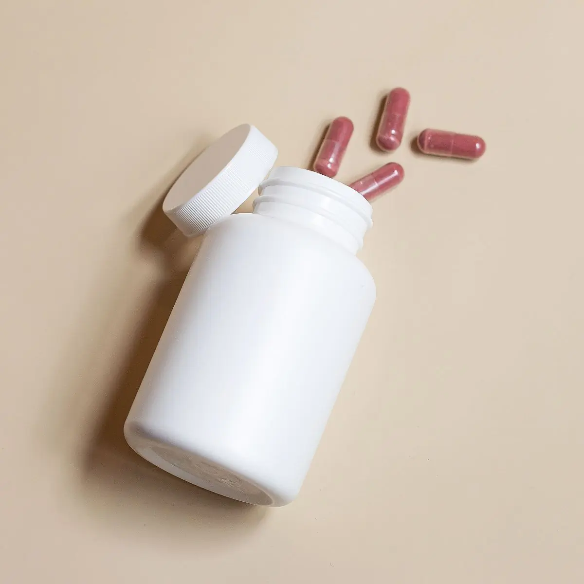 An uncapped white bottle photographed from above with capsules tumbling onto a beige surface