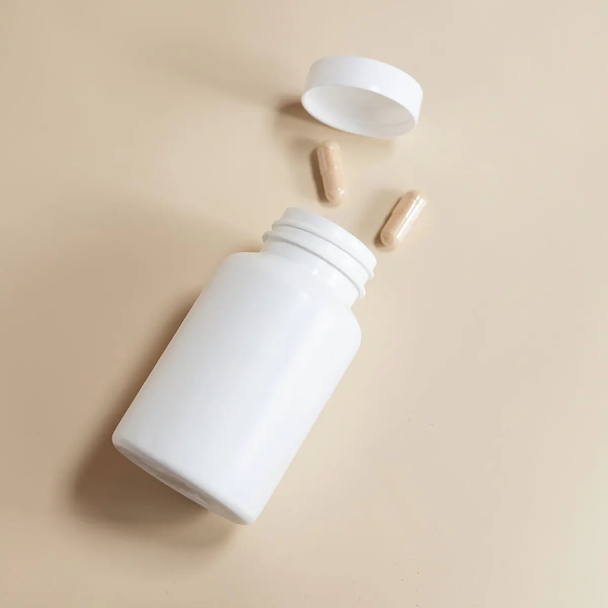 An open white supplement bottle lying on a beige surface with its cap off and two capsules alongside