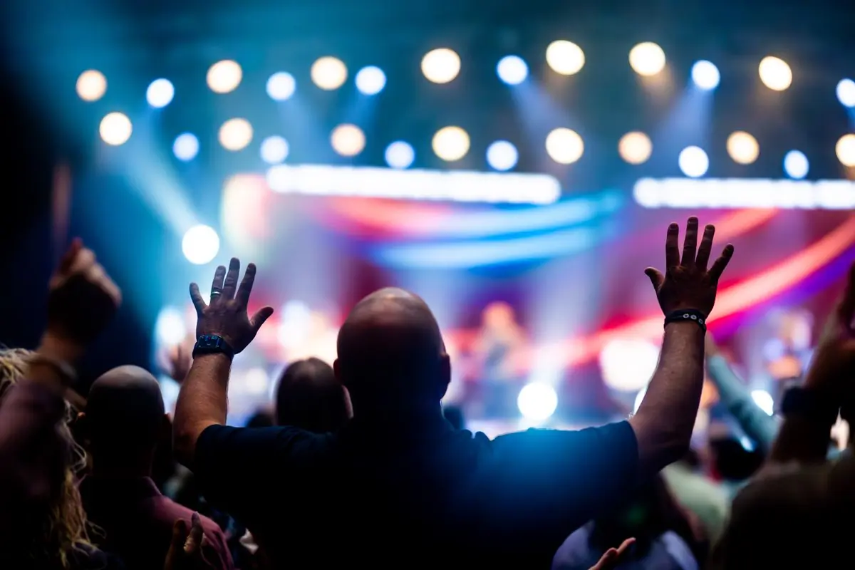 A brightly lit stage seen over the raised hands of an audience