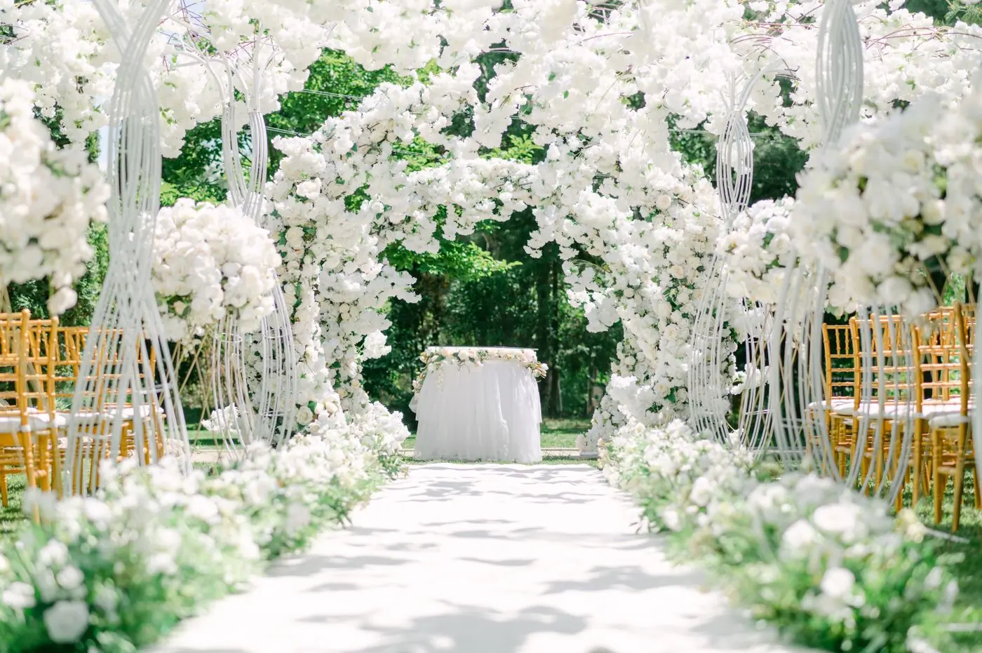 A garden aisle dressed with white blossom arches before a ceremony