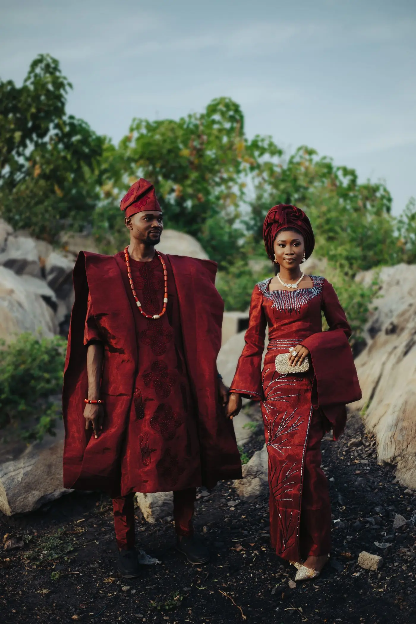 A couple in deep red traditional cloth and coral beads walking over rocks