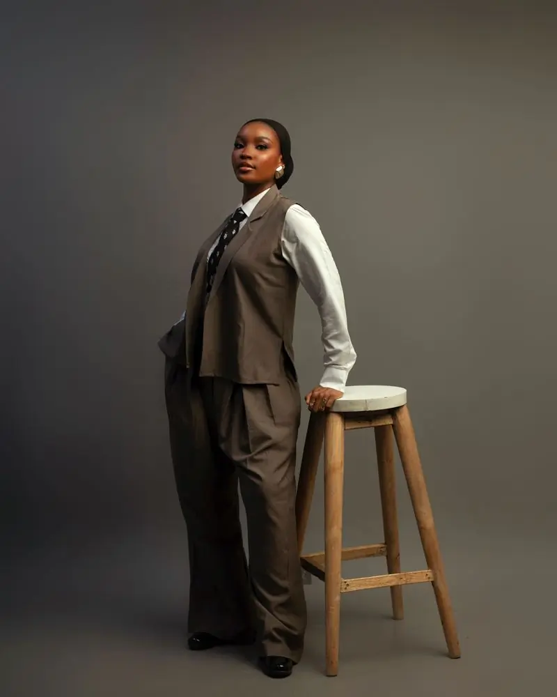A woman in a tailored grey suit standing beside a stool in a studio
