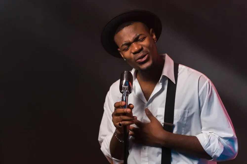A musician in a hat singing into a vintage microphone under a warm light