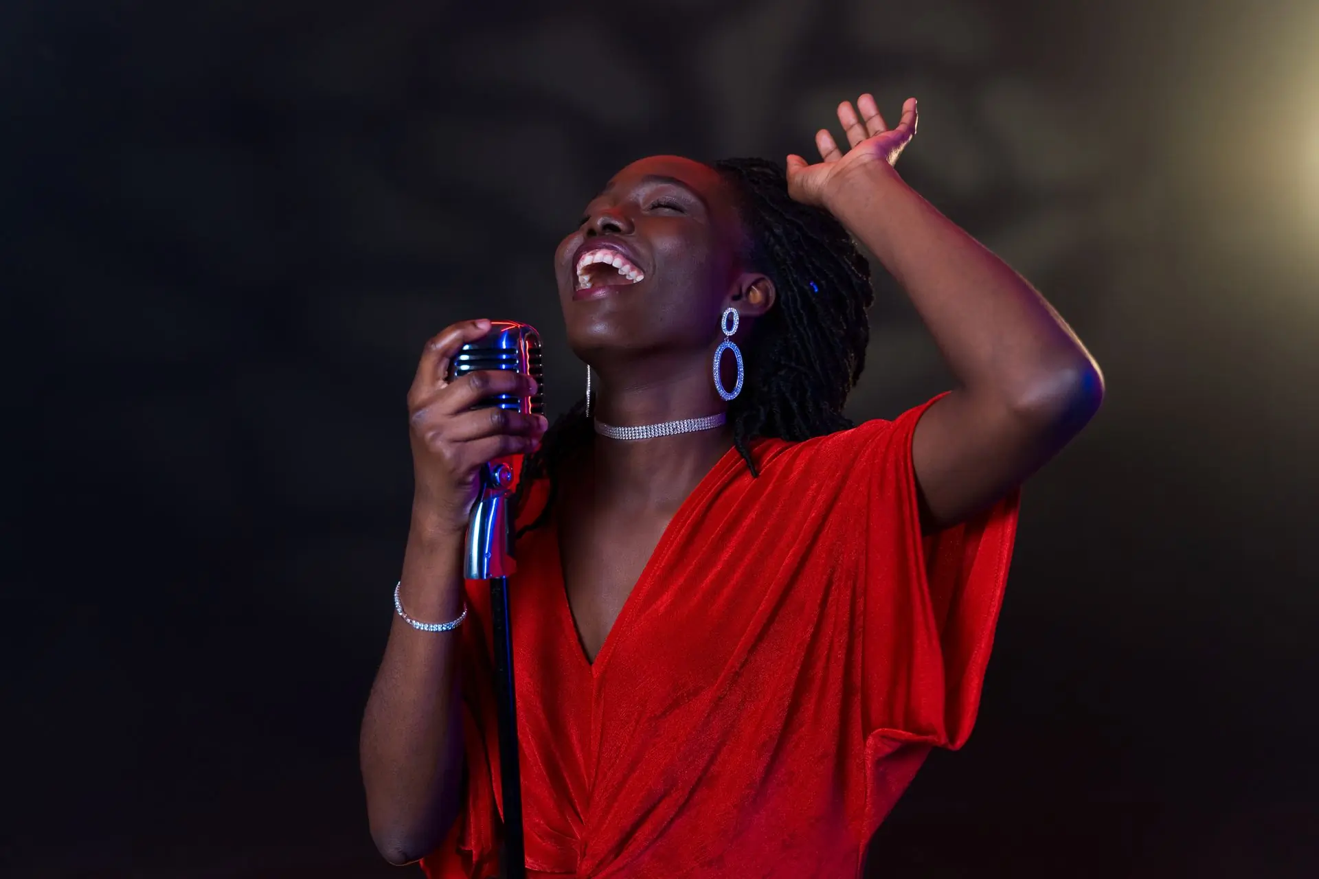A vocalist in a red dress singing into a chrome microphone under a single warm light