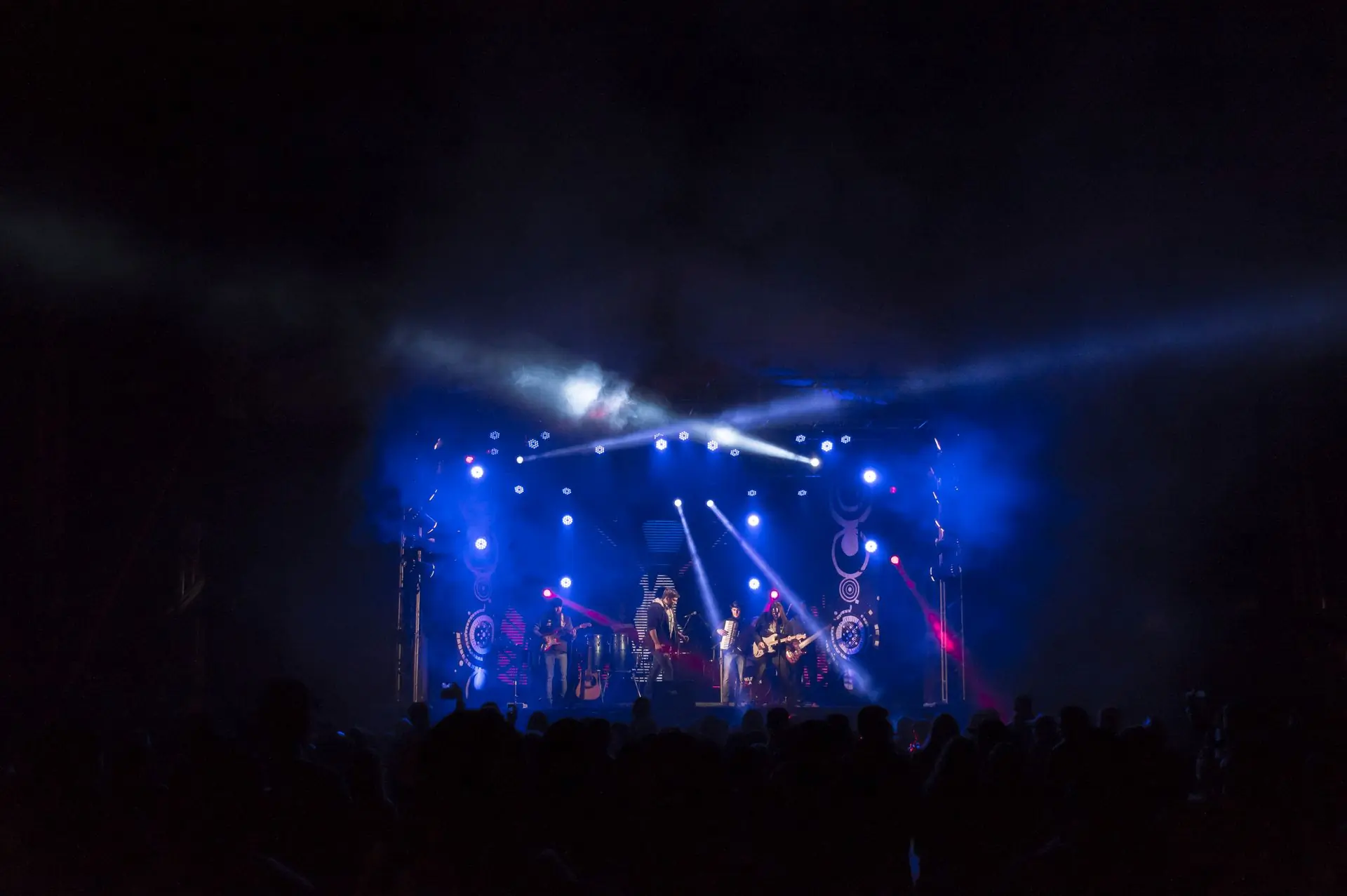 A band on a stage washed in blue light, seen over the heads of the crowd